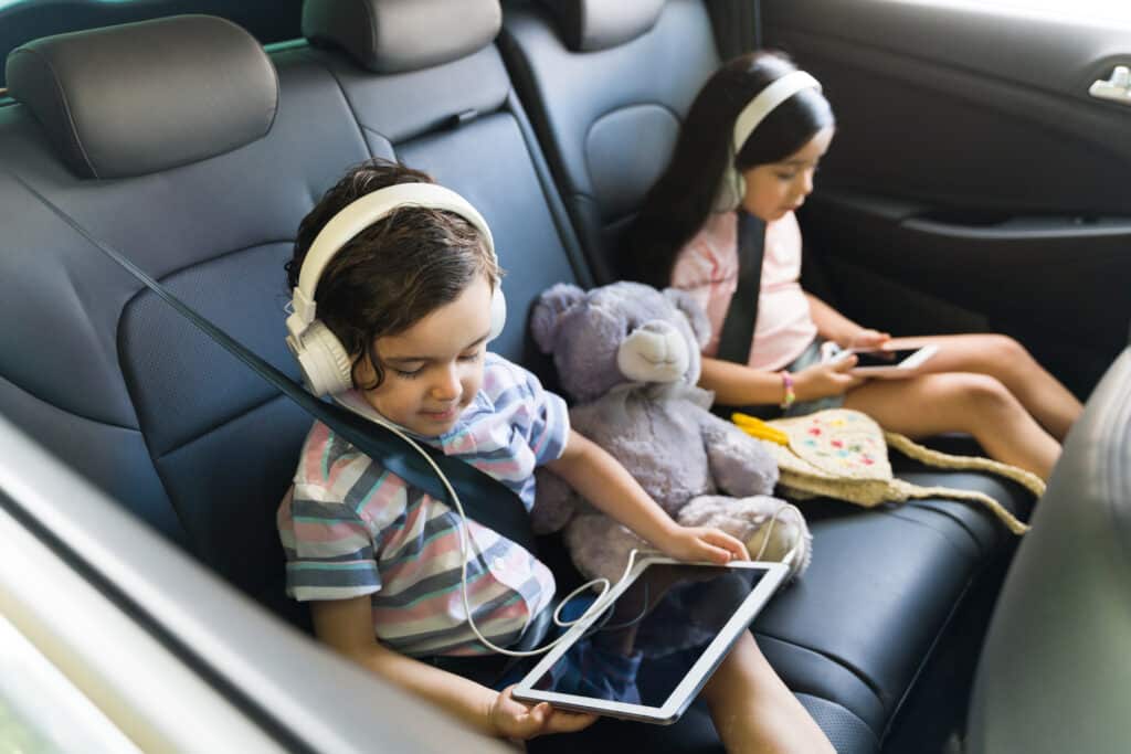 Deux enfants concentrés dans le siège arrière d'une voiture. Un garçon utilise une tablette avec un casque audio, une fille un smartphone.