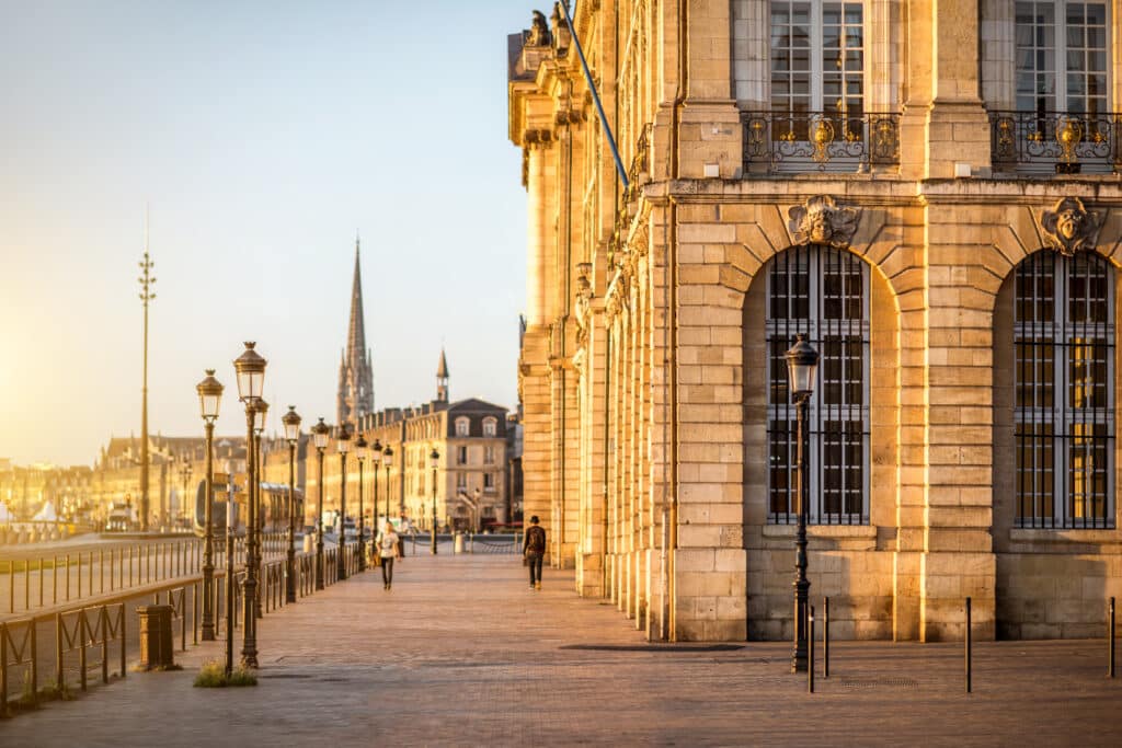Façade en pierre dorée à Bordeaux. Réverbères et quai longeant la Garonne au soleil couchant vers la flèche de Saint-Michel.